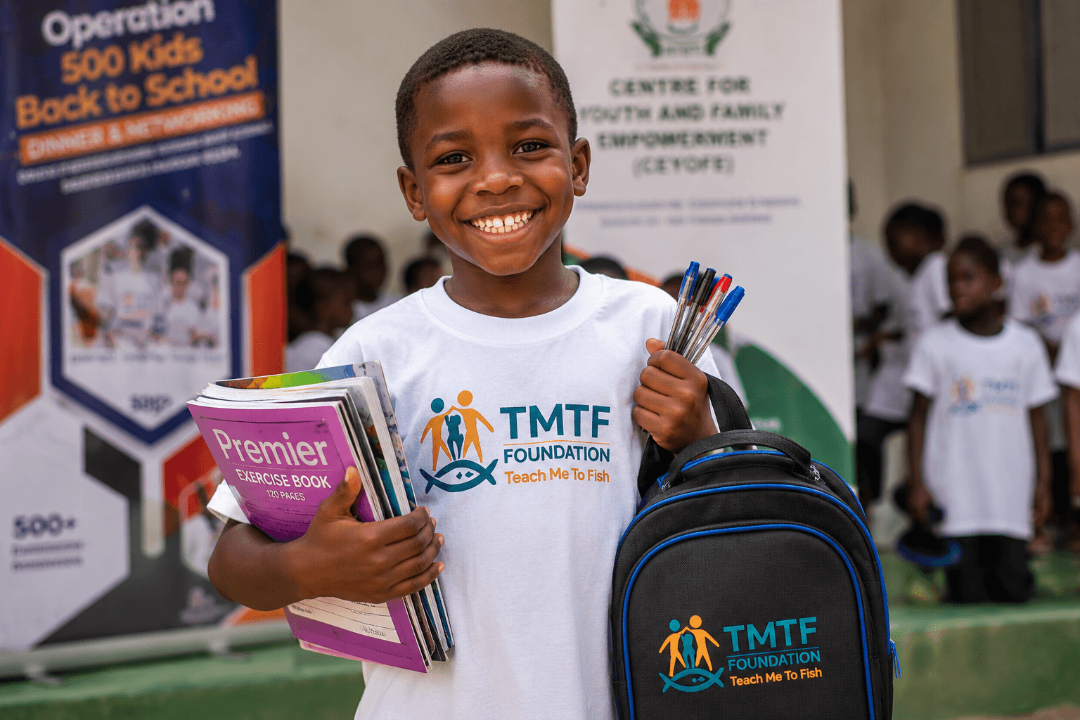 A boy in a Teach Me To Fish T-shirt smiling and holding a stack of exercise books, pens and a TMTF backpack