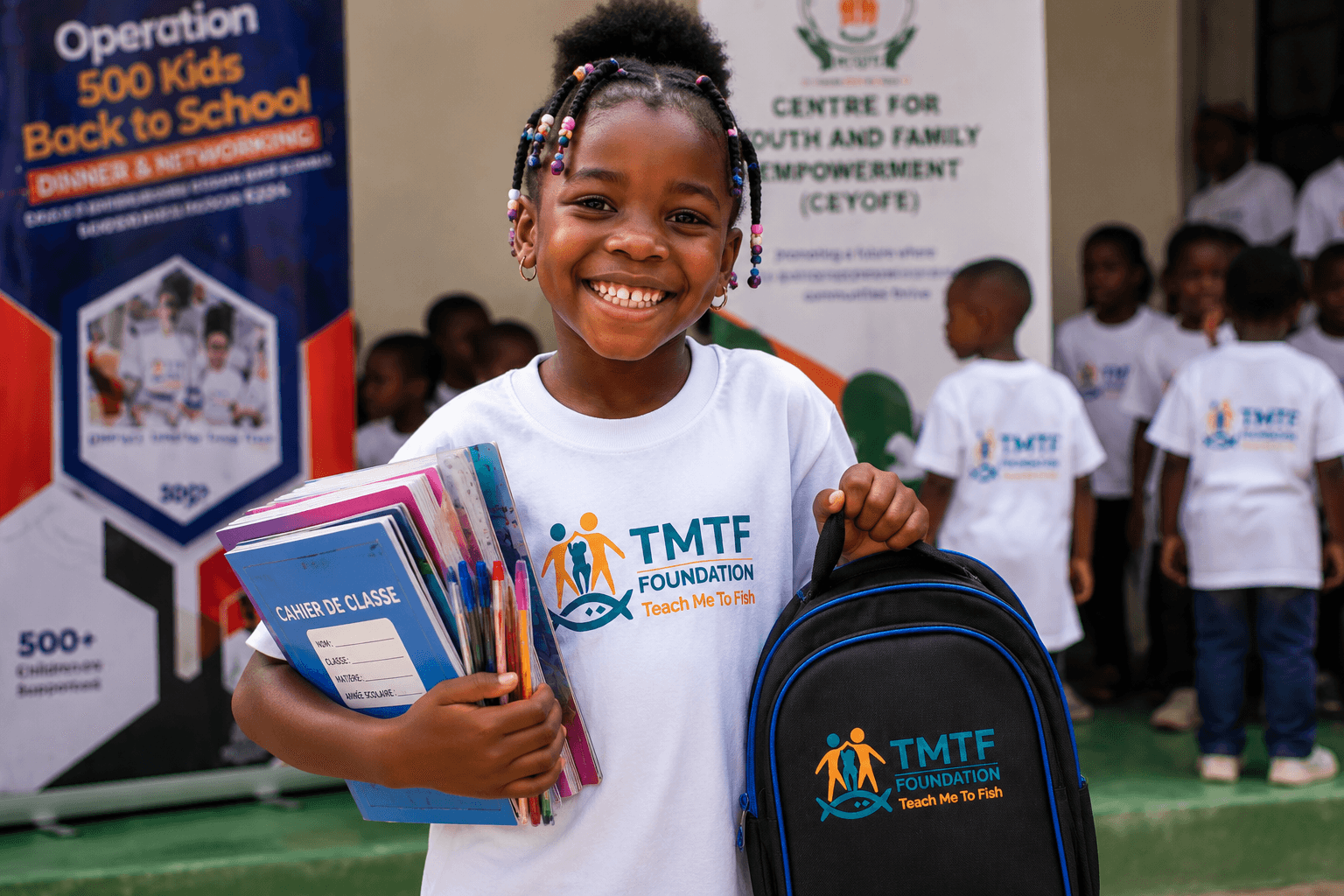 A girl in a Teach Me To Fish T-shirt smiling and holding her exercise books, pens and a TMTF backpack