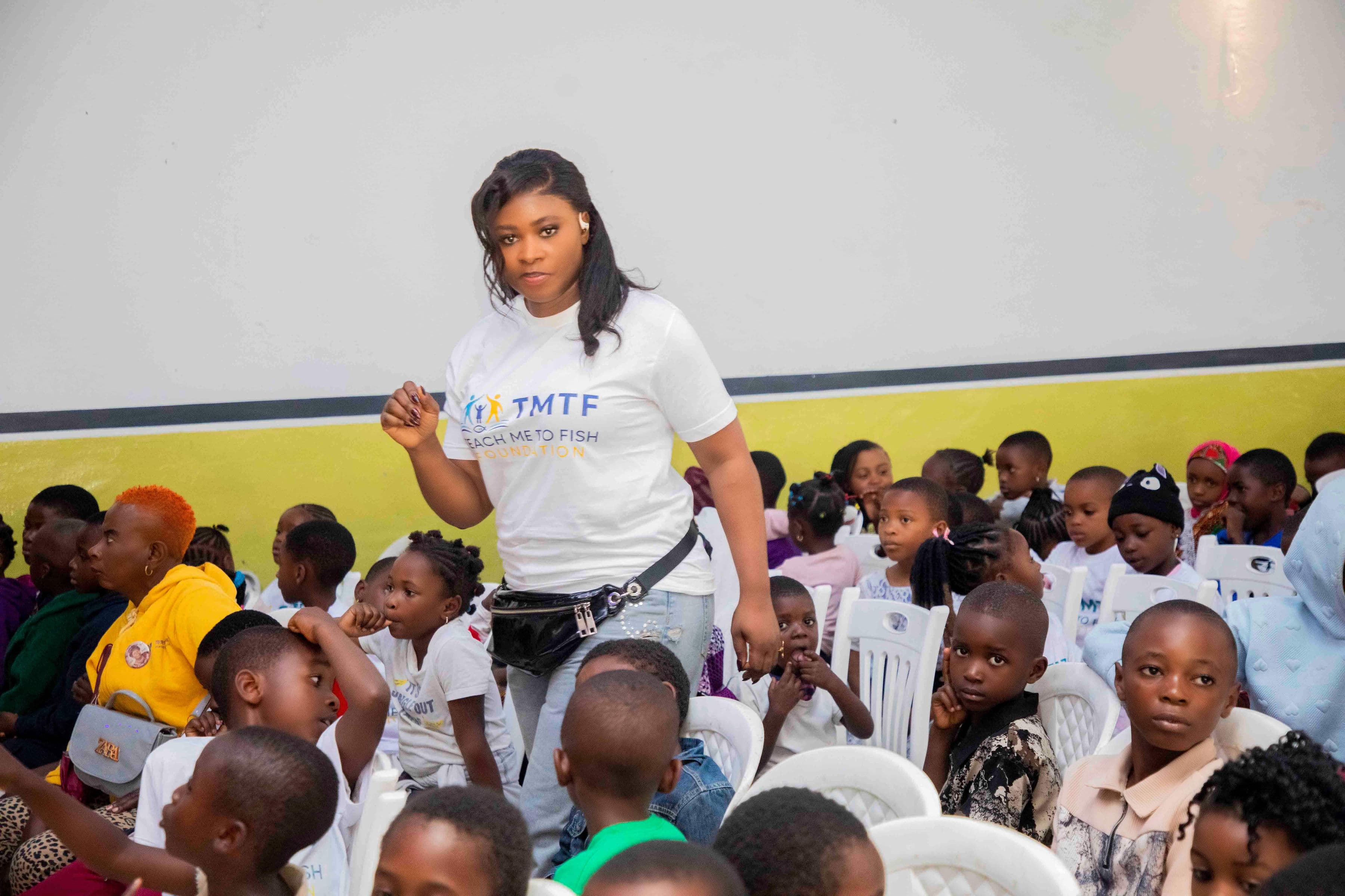 A woman in a Teach Me To Fish T-shirt standing in the aisle between rows of white plastic chairs filled with children and parents, against a white and yellow hall wall