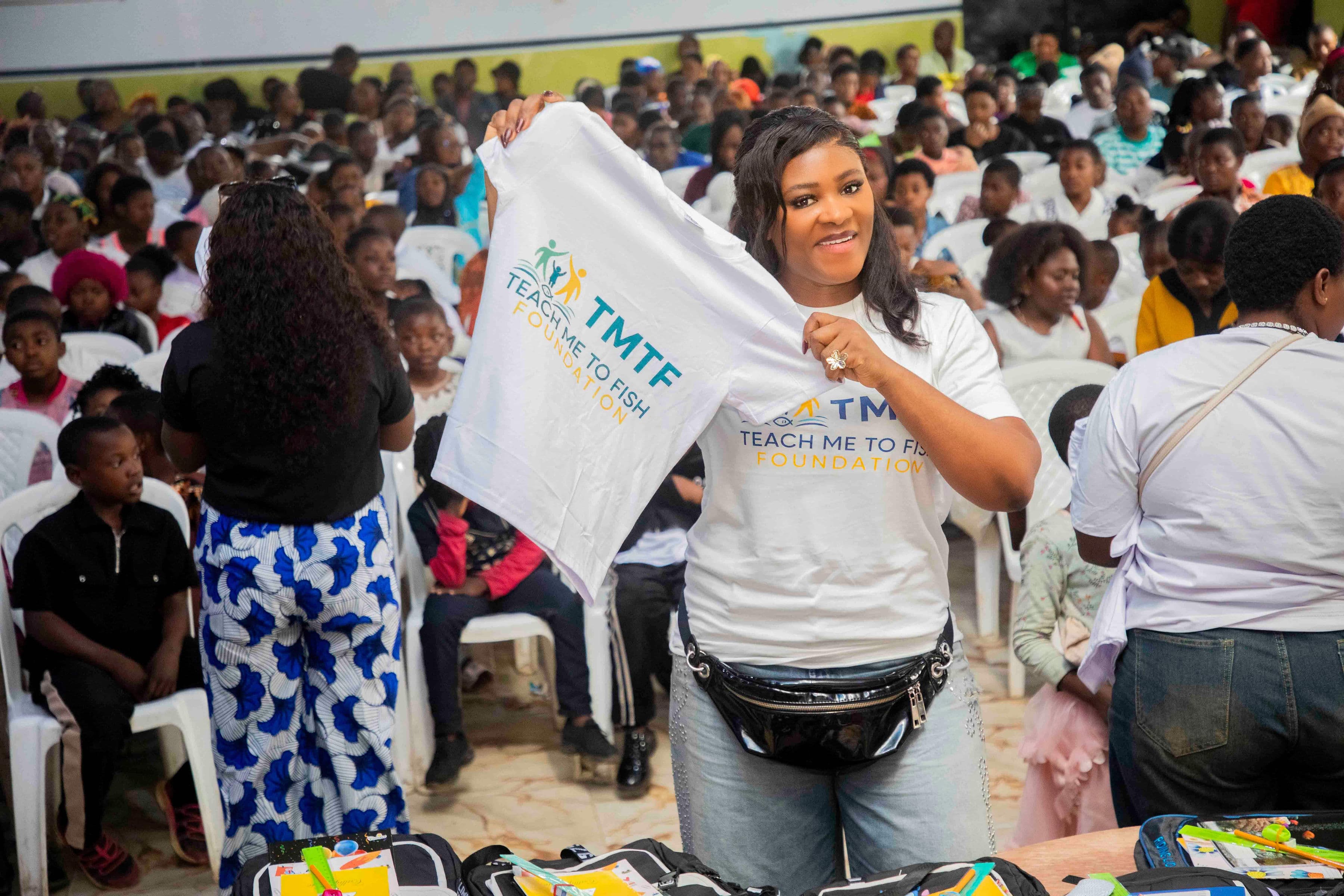 A woman in a Teach Me To Fish T-shirt holding up a second TMTF T-shirt to show a hall packed with several hundred seated children and parents, with opened backpacks full of stationery on a table in the foreground