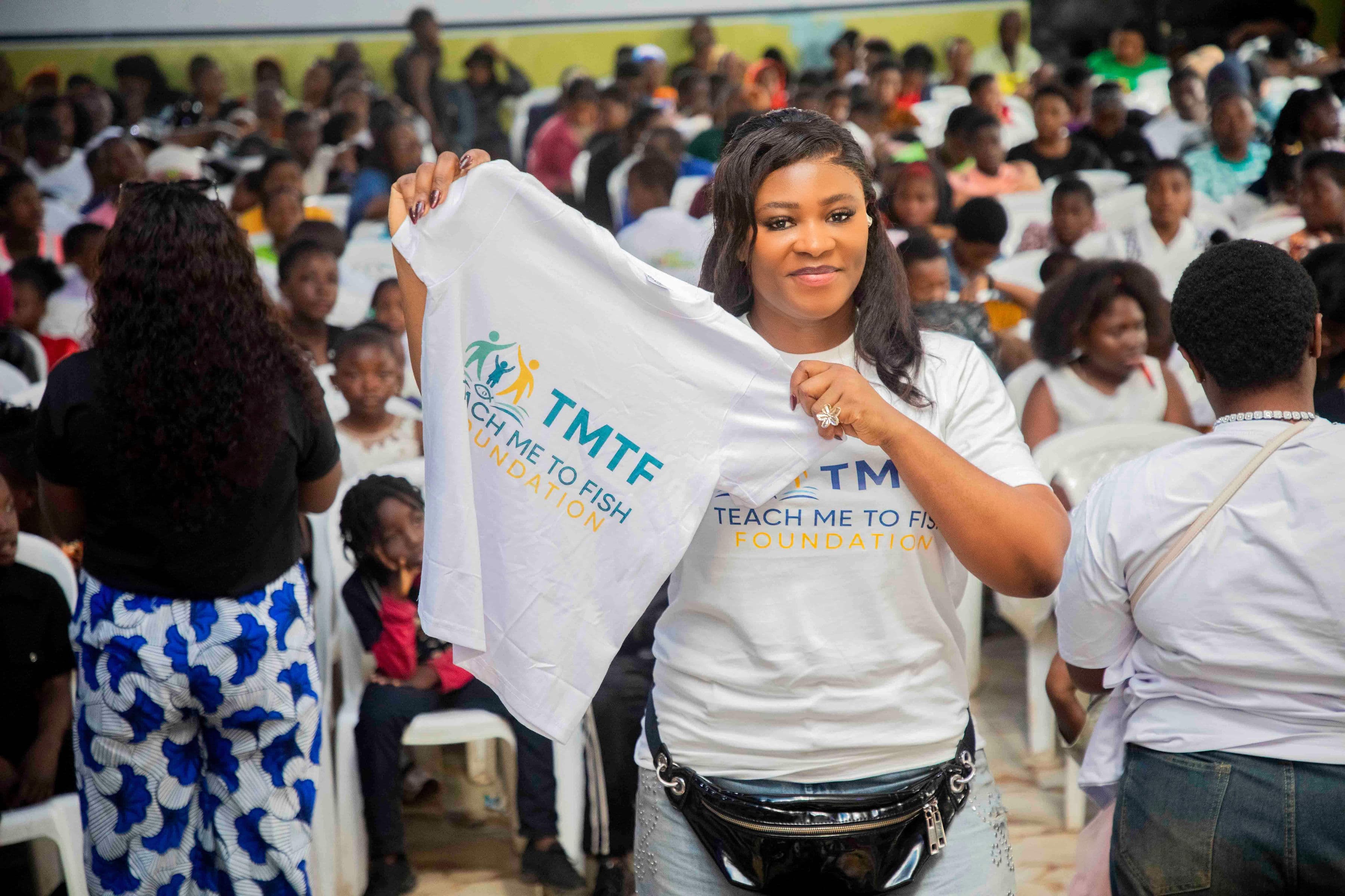 The same woman holding up a Teach Me To Fish T-shirt bearing the foundation's logo, turned towards the camera, with the crowded hall of families behind her