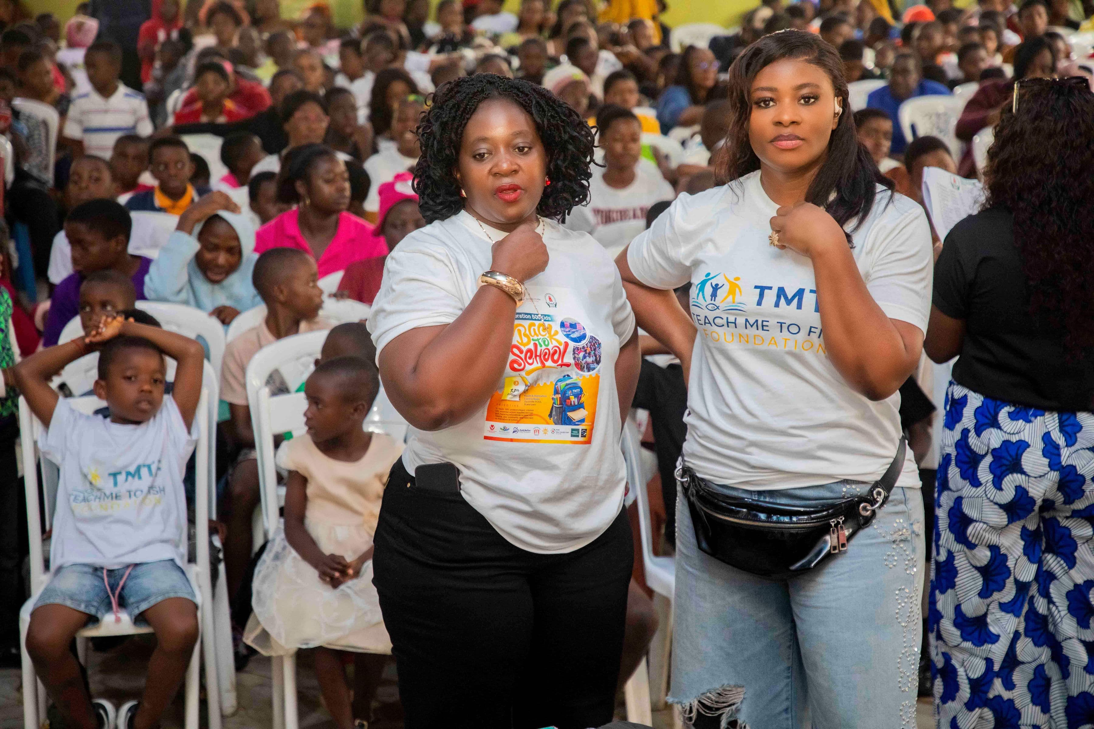 Two women standing side by side in the hall, one in a Back to School campaign T-shirt showing a backpack and partner logos, the other in a Teach Me To Fish shirt, with seated children and families around them