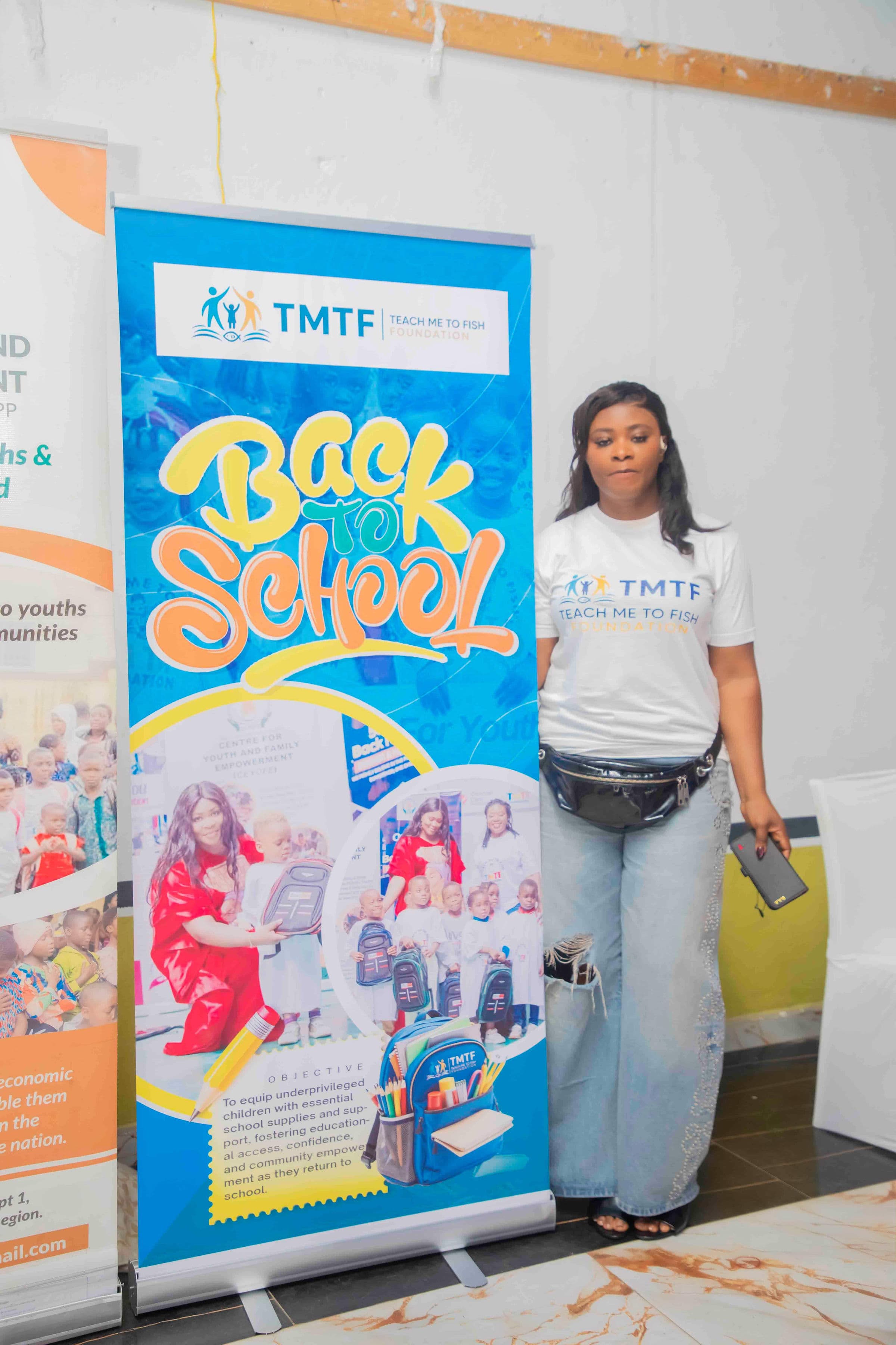 A woman in a Teach Me To Fish T-shirt standing beside a tall Back to School pull-up banner carrying the TMTF logo, photographs from previous events and the campaign's stated objective
