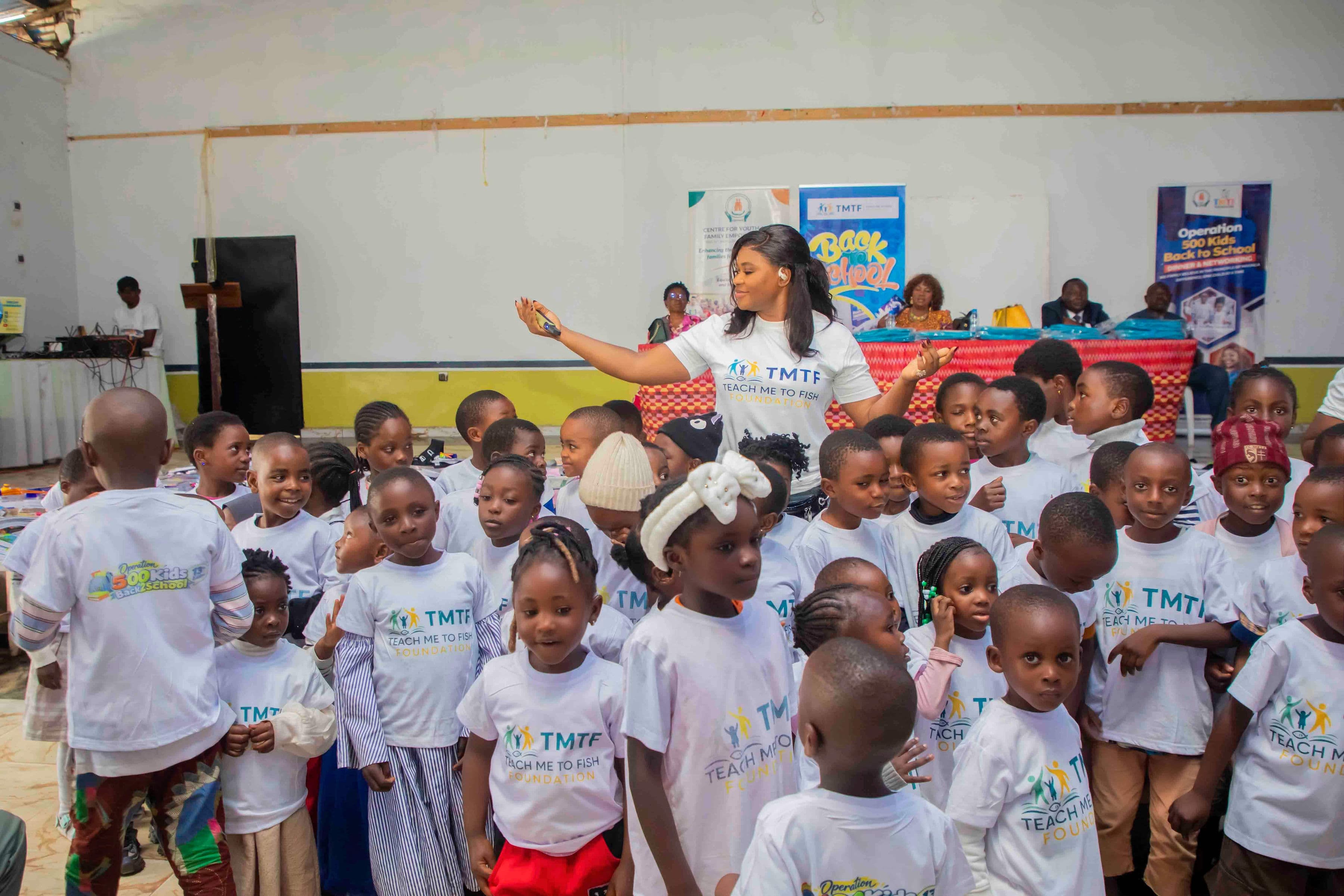 Several dozen children in Teach Me To Fish T-shirts gathered at the front of the hall, with a woman from the team standing among them with her arms open, and partner banners and a long table of packed bags behind