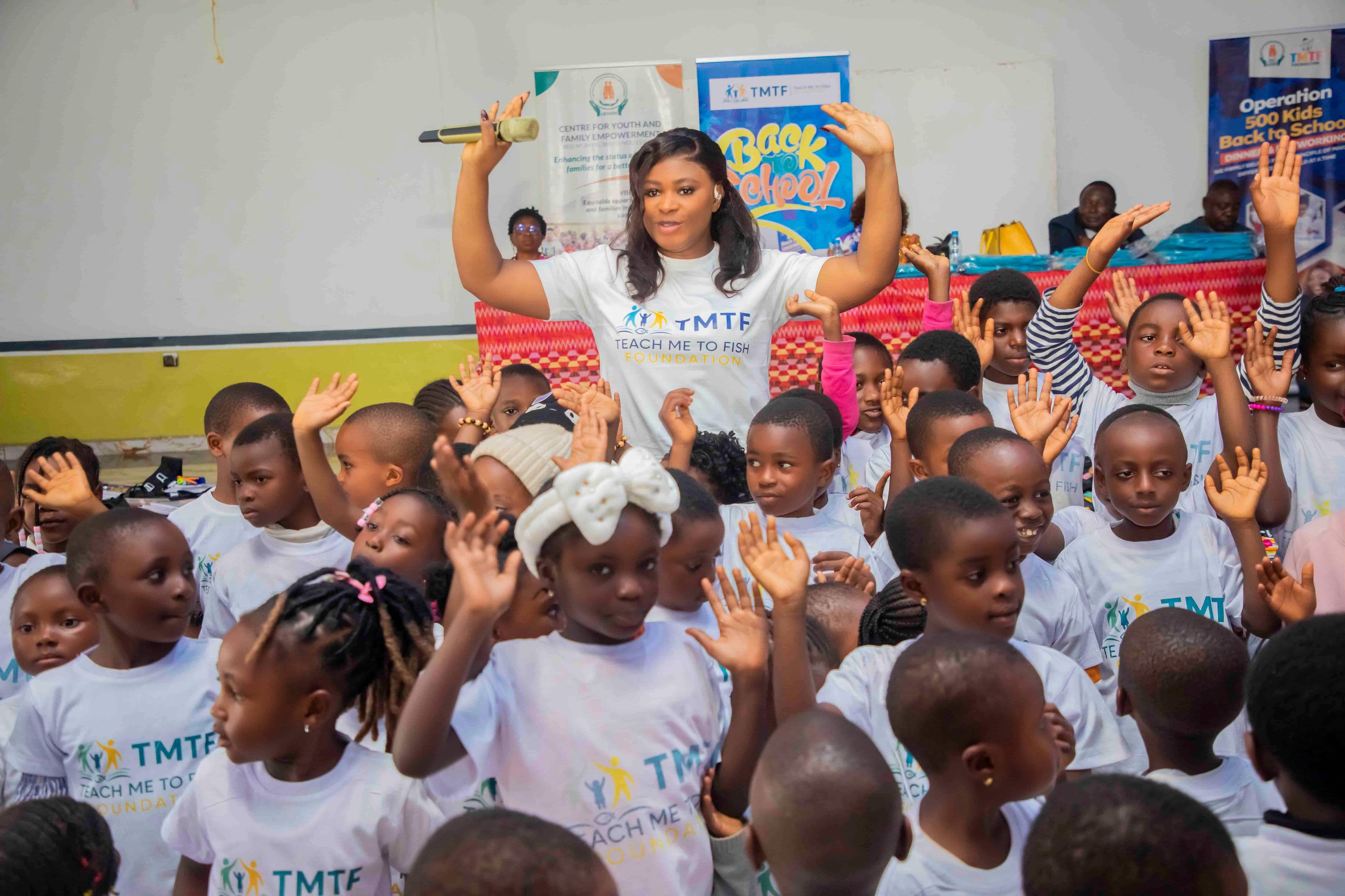 Children in Teach Me To Fish T-shirts with their hands in the air, answering a woman who stands among them holding a microphone with both arms raised, in front of Back to School and Operation 500 Kids banners