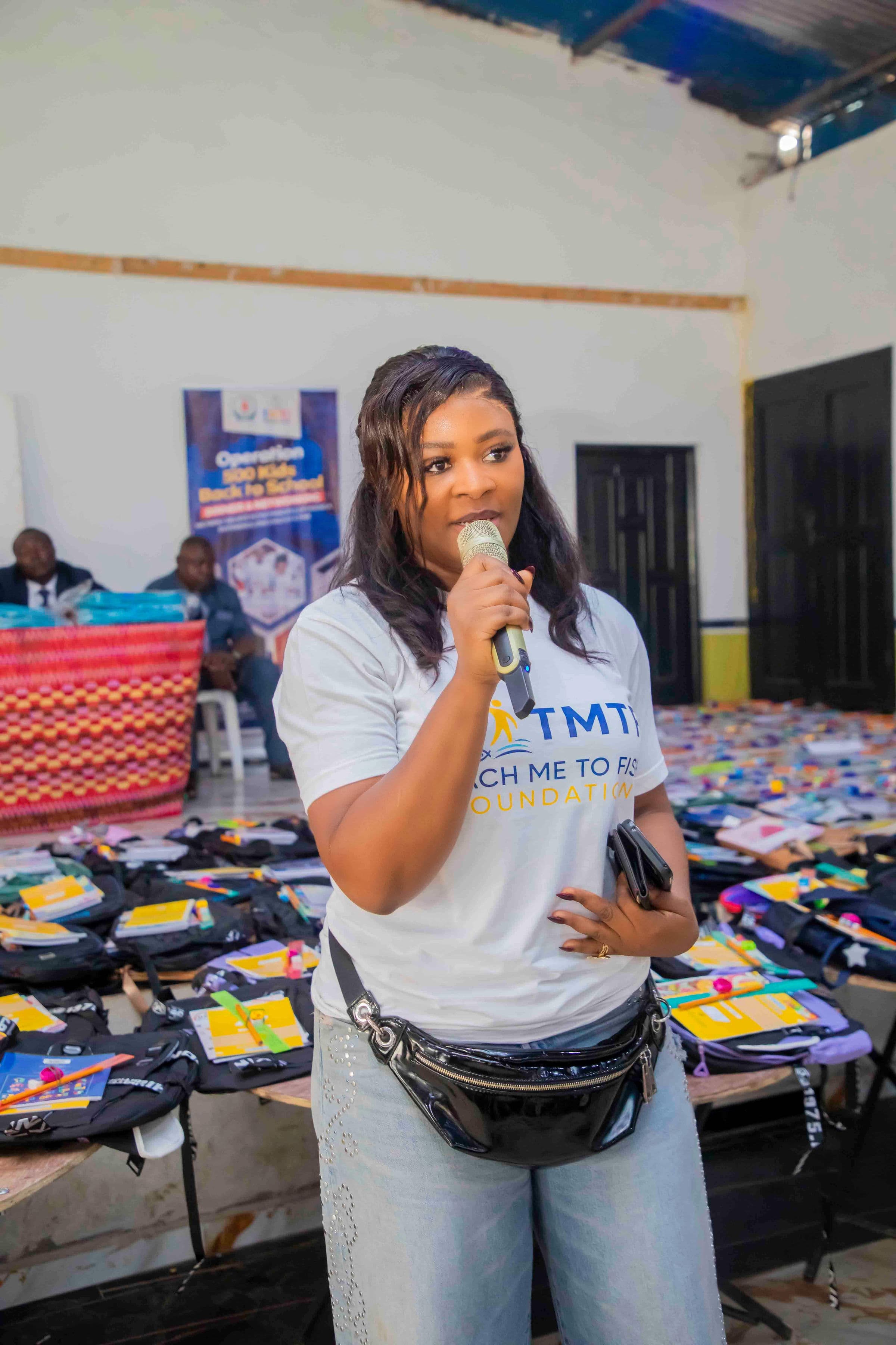 A woman in a Teach Me To Fish T-shirt speaking into a microphone, with long tables behind her carrying dozens of open backpacks filled with exercise books, pencils and rulers