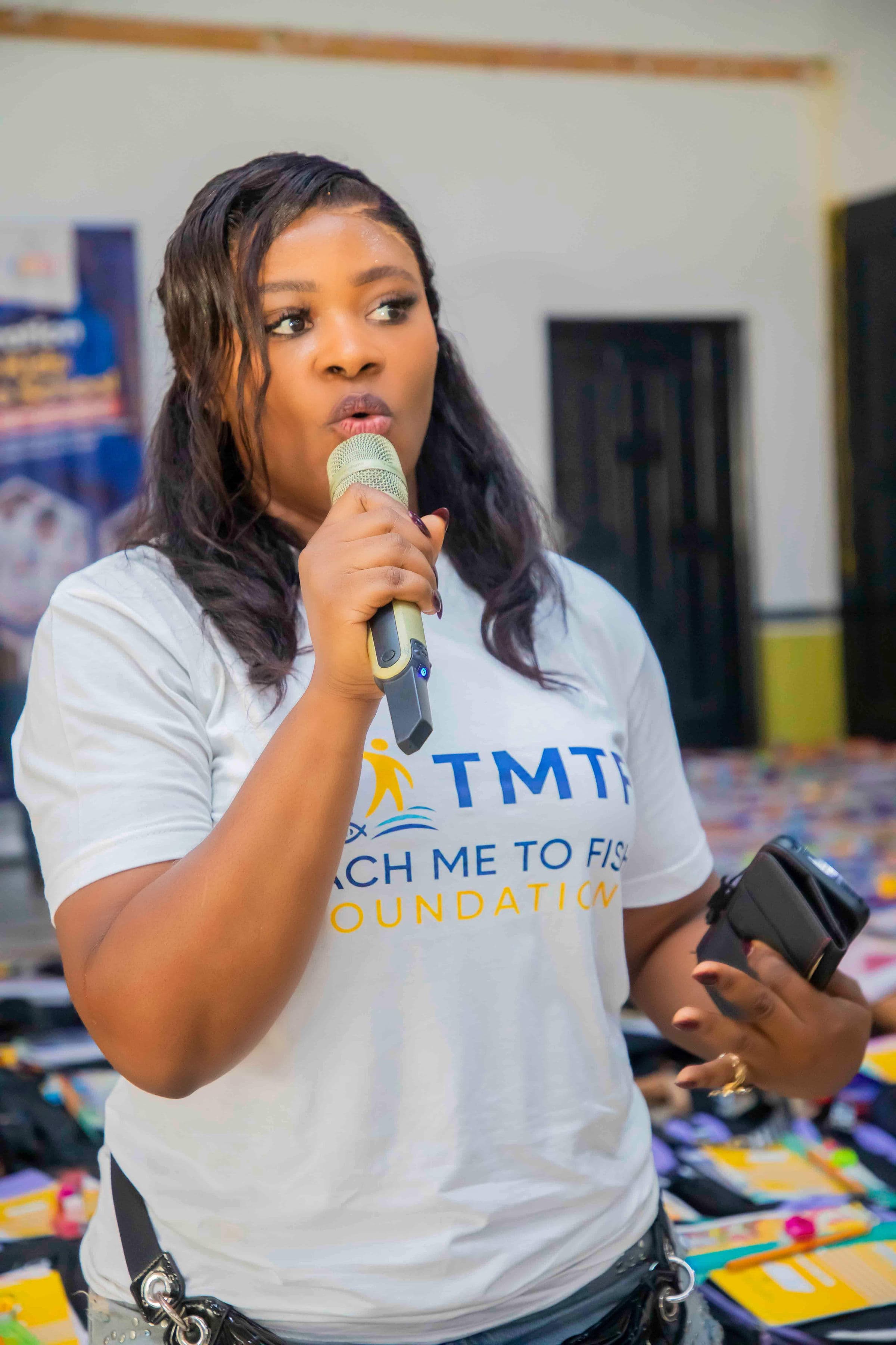Close portrait of the same woman speaking into a microphone in the hall, with the tables of packed backpacks out of focus behind her