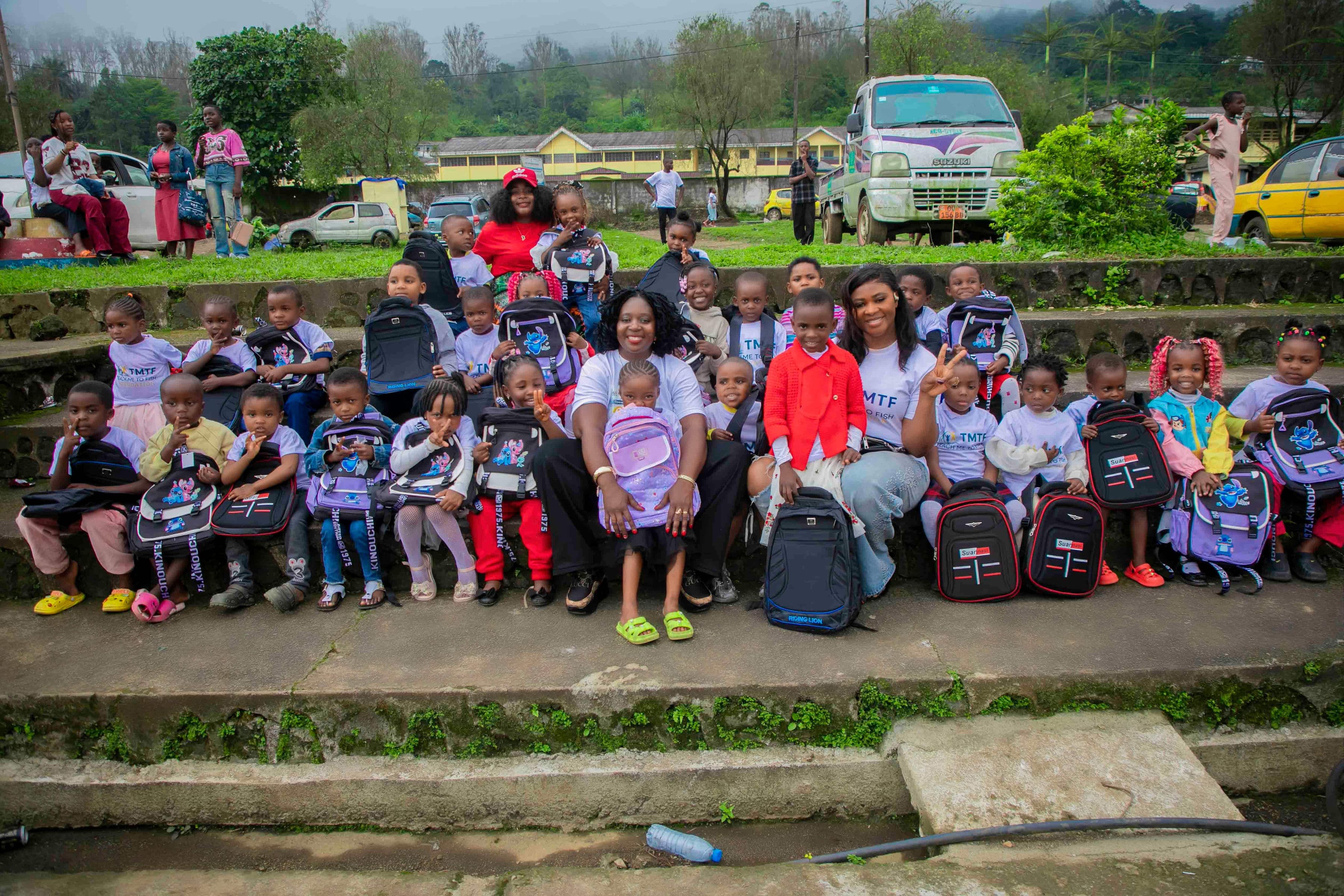 About thirty young children in Teach Me To Fish T-shirts sitting in rows on wide concrete steps outdoors, each with a new backpack beside them, with three women from the team among them and green hills, a school building and parked cars behind