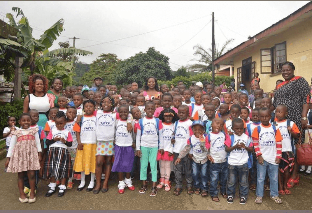 A large group of children in TMTF Foundation T-shirts standing outdoors with staff, in front of a building with green hills behind