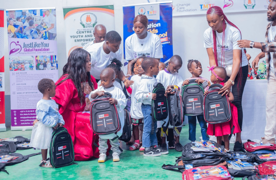A group of young children each holding a new school backpack, with volunteers in JustLikeYou Global Foundation shirts helping them, and partner banners behind