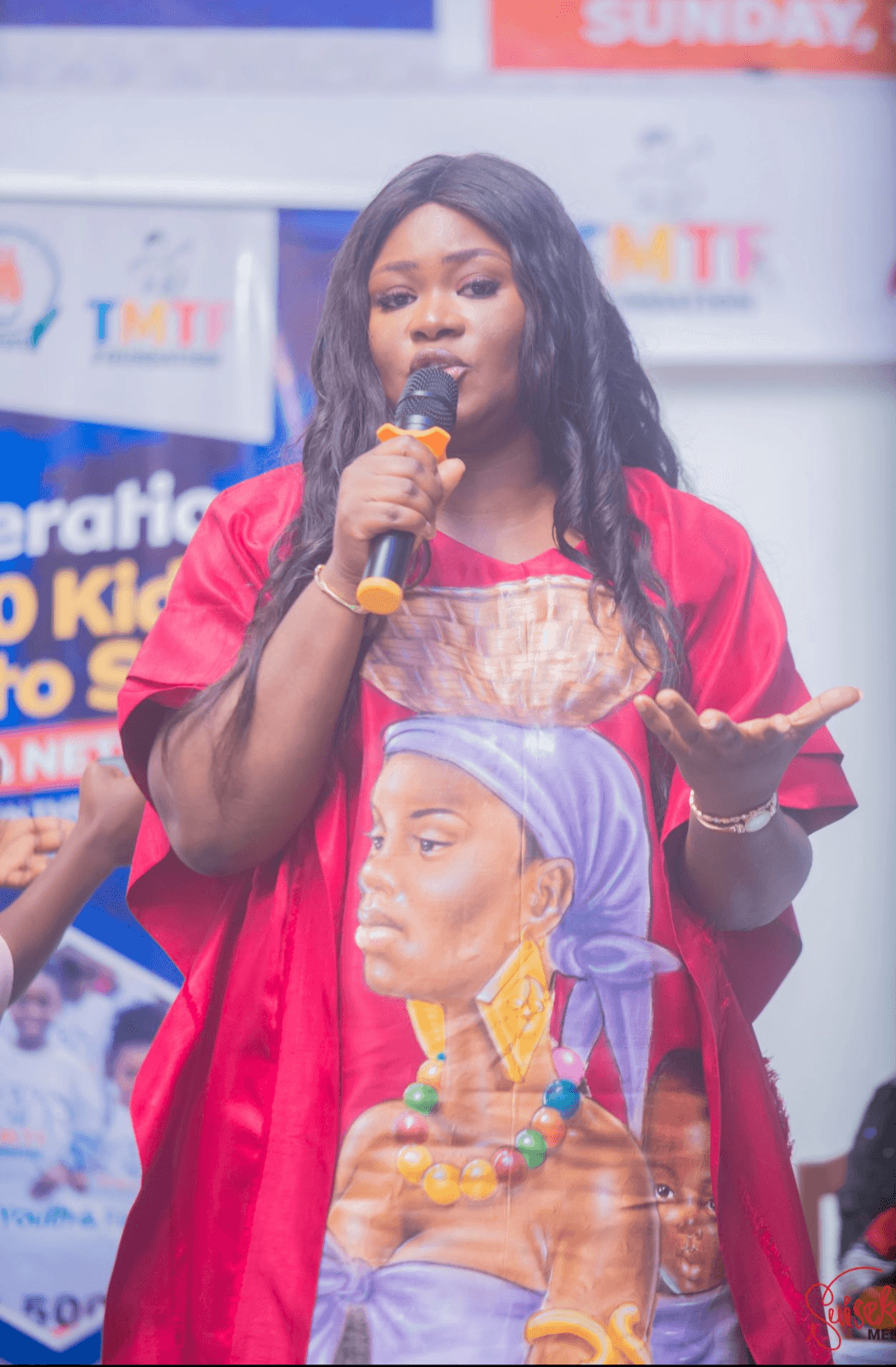 TMTF founder Dorothy Akoh-Arrey speaking into a microphone at a Back to School event, in front of TMTF and Operation 500 Kids banners