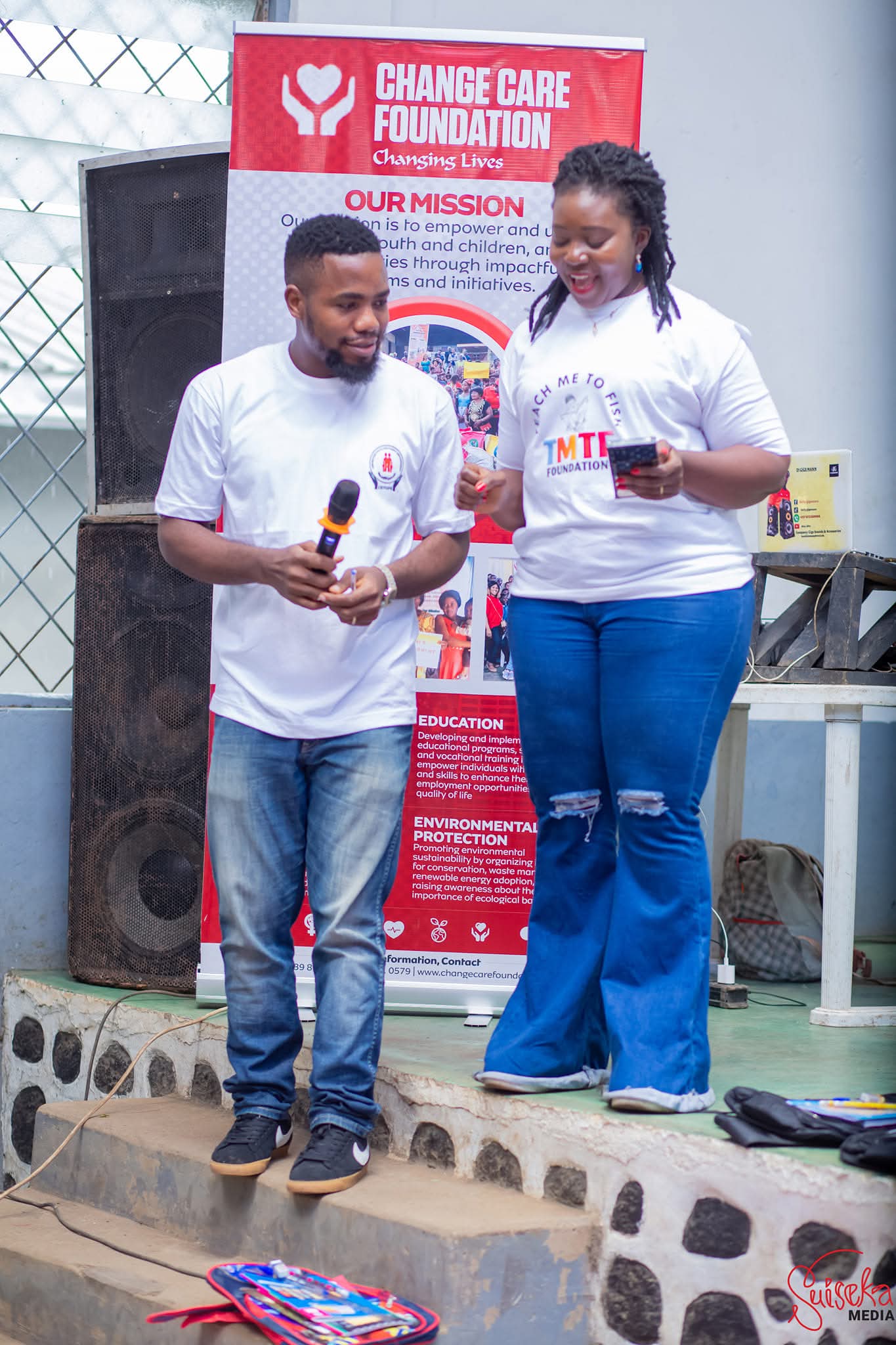 A CEYOFE team member holding a microphone on stage beside a colleague in a Teach Me To Fish T-shirt, in front of a partner banner, with a packed school bag on the step in front of them
