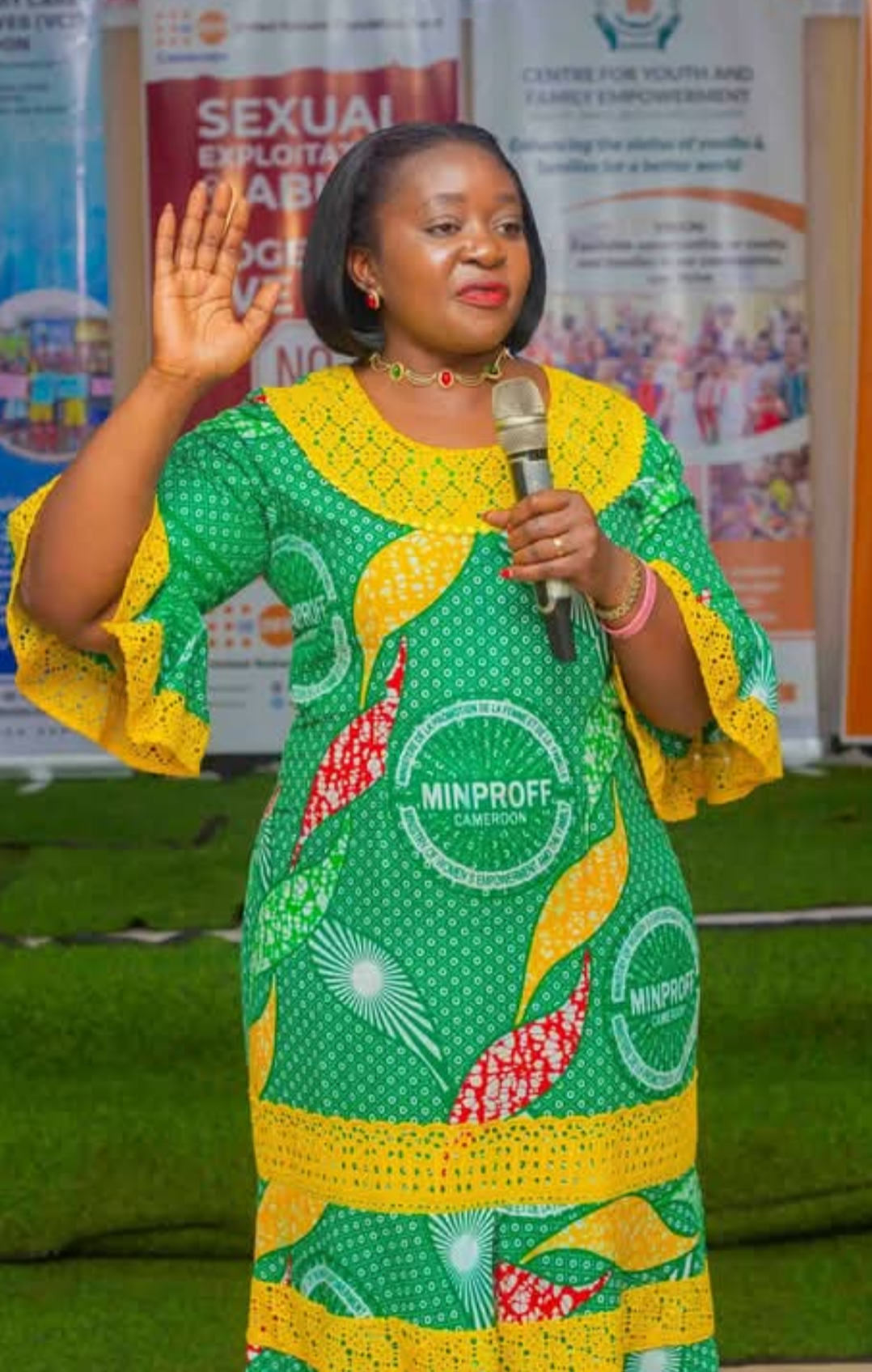 Eteki Stella Dopgima speaking into a microphone with one hand raised, wearing a green and yellow MINPROFF print dress, in front of CEYOFE and women's empowerment banners
