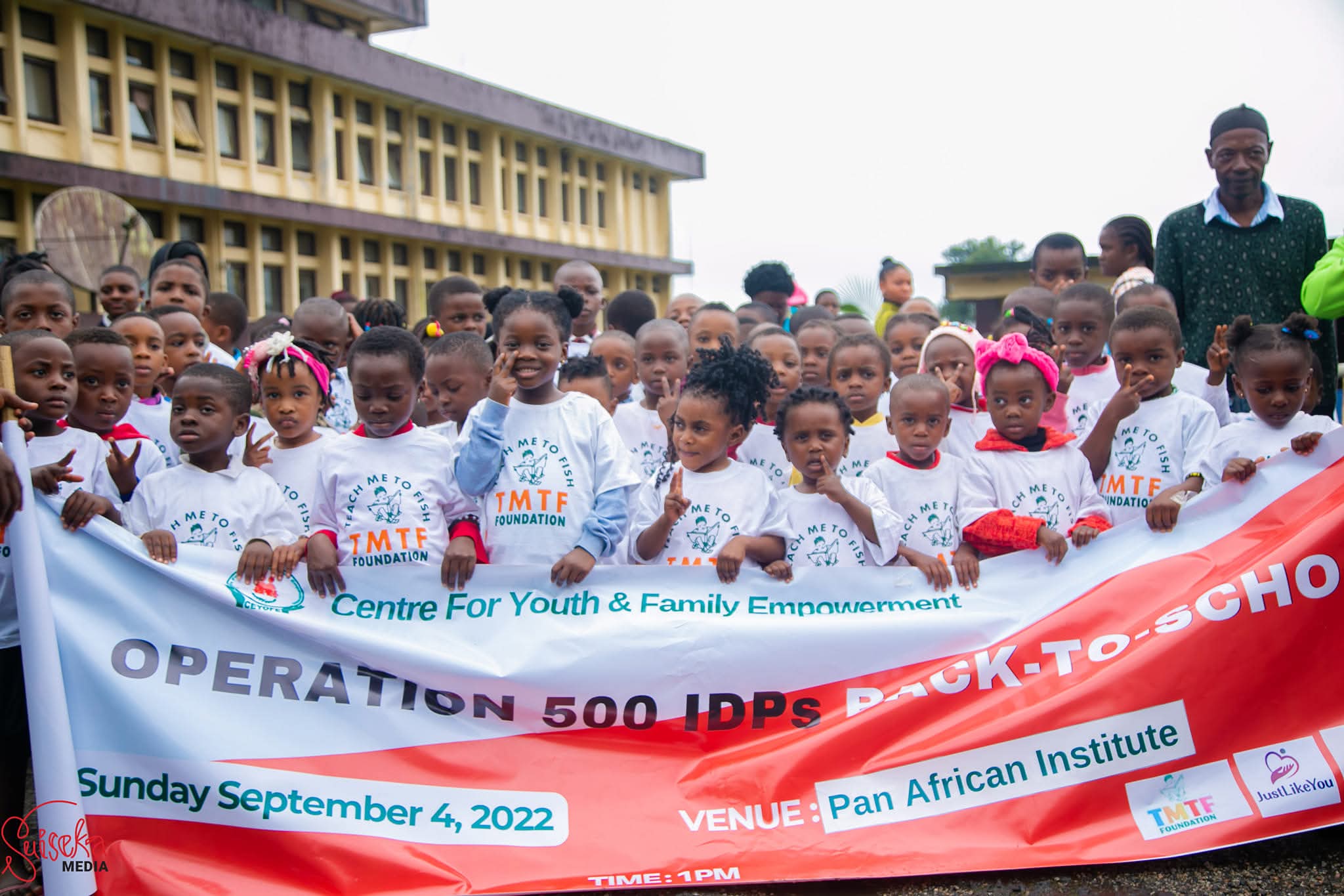 Dozens of young children in Teach Me To Fish T-shirts standing behind a Back to School banner outside the Pan African Institute in Buea