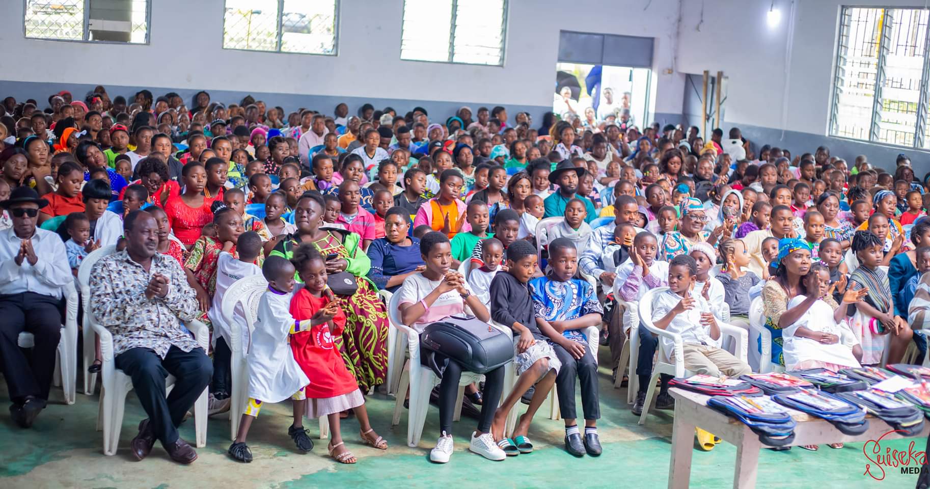 A hall filled with several hundred seated children and parents at a Back to School event, applauding, with tables of packed school bags along the right-hand side