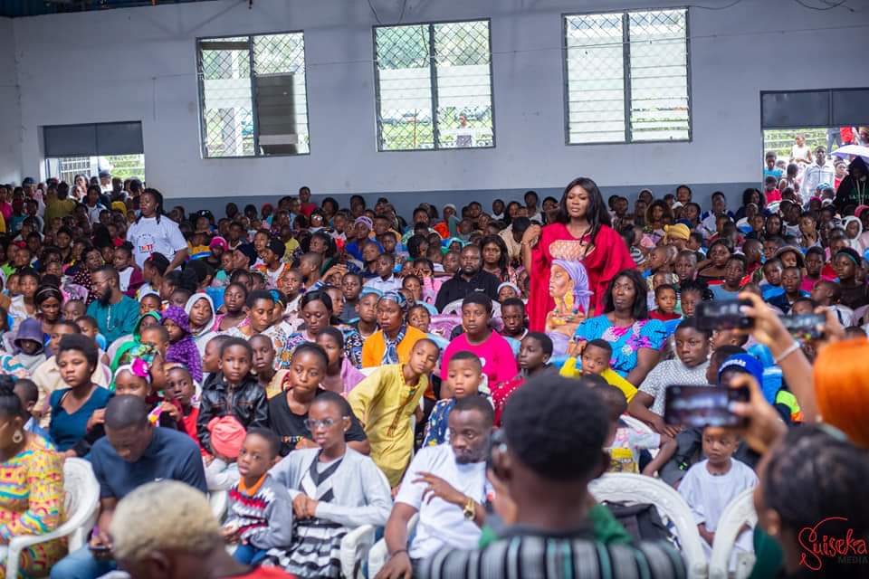 TMTF founder Dorothy Akoh-Arrey standing in a red gown in the middle of a crowded hall of seated children and families at a Back to School event