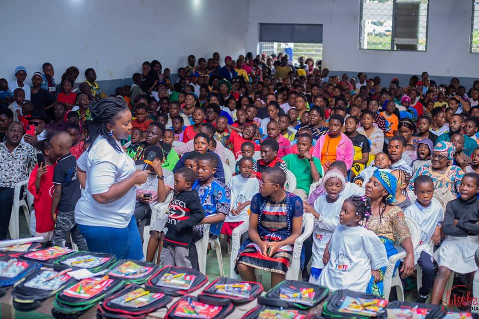 A TMTF volunteer with a microphone talking to children at the front of a packed hall, with rows of open backpacks full of pens and exercise books on the table in front