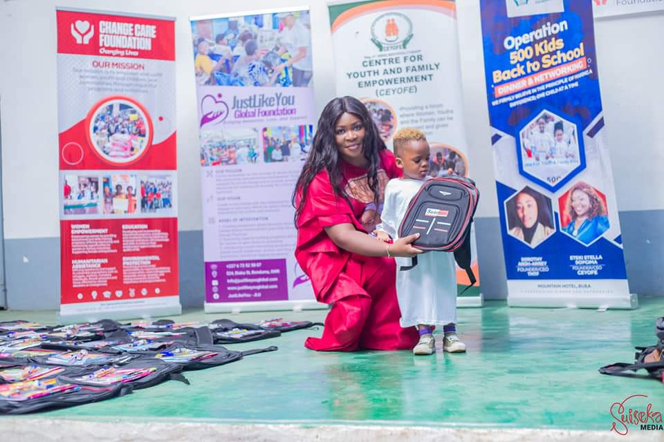 TMTF founder Dorothy Akoh-Arrey kneeling to hand a TMTF backpack to a small child, in front of partner banners, with dozens of packed bags laid out on the floor