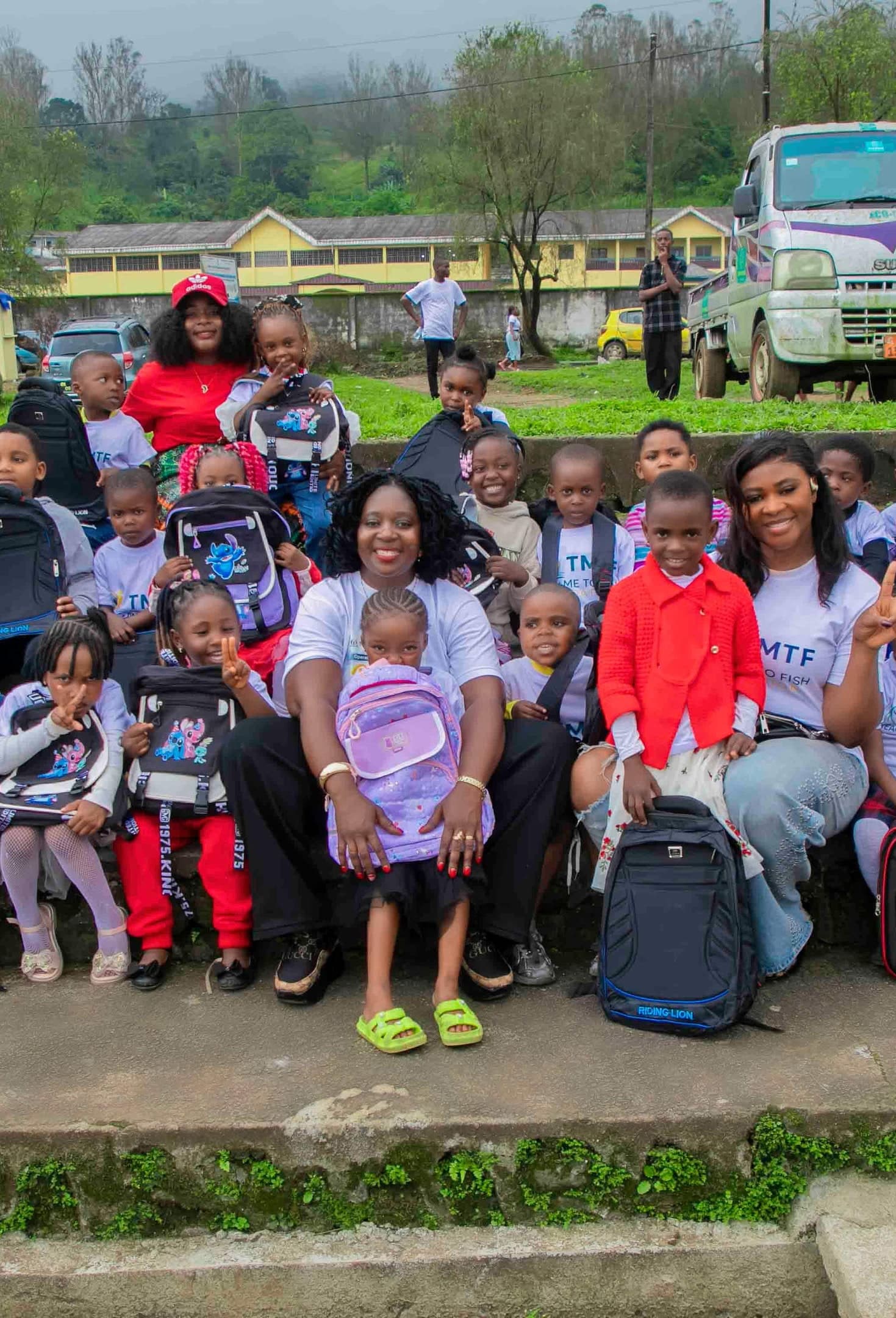 About twenty young children in Teach Me To Fish T-shirts sitting together on wide concrete steps outdoors, each with a new school backpack, with two women from the team among them and a school building and green hills behind